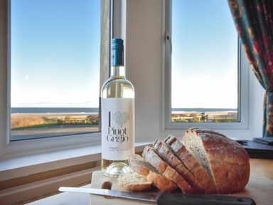 A sliced loaf of bread and a bottle of white wine on a table by a window with a view of the beach at Claremount in Alnmouth