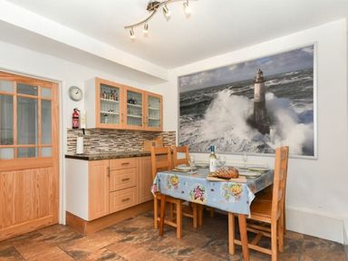 A dining area with a wooden table covered with a tablecloth and wooden chairs near cabinets and a large framed picture of a lighthouse at Claremount in Alnmouth