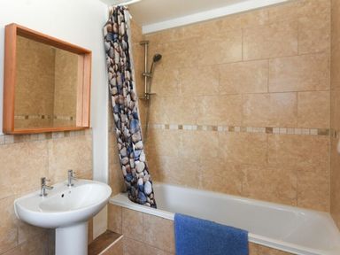 A bathroom with a bathtub shower combination a stone patterned shower curtain a blue towel on the tub and a sink with mirror above at Claremount in Alnmouth