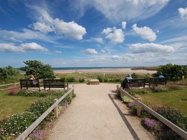 A path leading to benches overlooking a beach with sand dunes and the sea under a blue sky at Claremount in Alnmouth