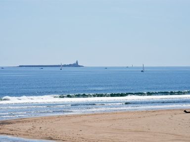 A sandy beach with waves and sailboats on the ocean with a lighthouse in the distance at Claremount in Alnmouth