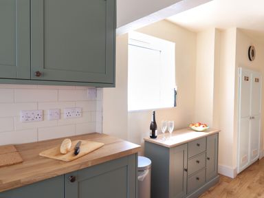 A kitchen with green cabinets wooden countertops a cutting board with bread and knife a window a sideboard with a bottle glasses and a bowl of fruit at 30 Fleet Street in Beaminster