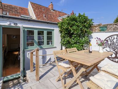 An outdoor patio with a wooden table and chairs with cushions a bottle of wine and glasses a flowering plant and green shrubbery at 30 Fleet Street in Beaminster