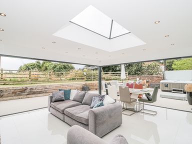 A living area with a grey sofa and dining table surrounded by large glass doors opening to an outdoor space with a hot tub at Orchard Leigh in North Petherton