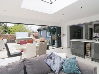 A modern open plan kitchen and dining area with a glass dining table and multicolored chairs with a sofa and cushions outside seating and a hot tub visible at Orchard Leigh in North Petherton