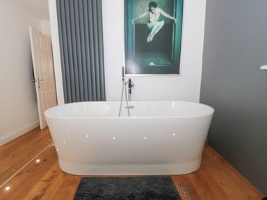 A bathroom with a white freestanding bathtub dark floor mat wooden floor and an artwork on the wall at Orchard Leigh in North Petherton