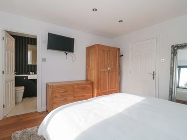 A bedroom with a white bedspread wooden wardrobe and dresser a wall-mounted TV an open door to a bathroom and a large framed mirror at Orchard Leigh in North Petherton