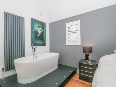 A bedroom with a bathtub on a raised platform a nightstand with a lamp and a glass a framed picture and a window with shutters at Orchard Leigh in North Petherton