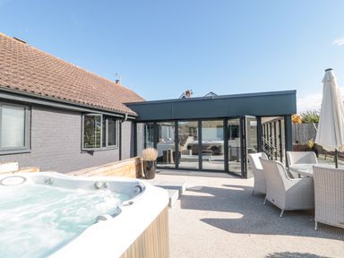An outdoor patio area with a hot tub, wicker chairs around a table, and a building with large glass doors at Orchard Leigh in North Petherton
