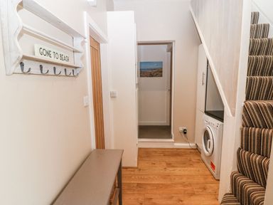 A hallway with wooden floor a bench a coat rack saying gone to beach a washing machine under stairs and a striped carpeted staircase at Victoria House in Rhosneigr