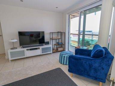 A living room with a blue armchair a striped pouf white tv stand with a television and a glass door leading to a balcony at Manhattan Beach in Castletown Portland