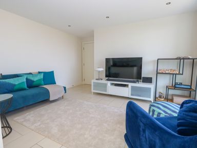 A living room with a blue sofa, television on a white stand, and a metal shelf with decorative items at Manhattan Beach in Castletown Portland