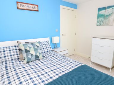 A bedroom with a bed with checkered bedding pillows and a blue accent wall with a sun sand surf sign at Manhattan Beach in Castletown Portland