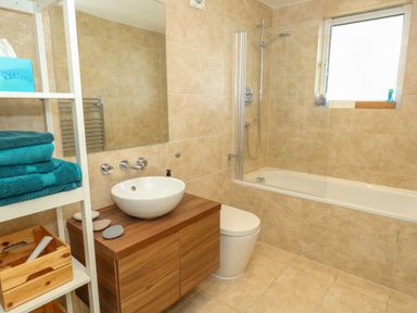 A bathroom with a bathtub shower combination a wooden vanity with a round sink a toilet and a white shelving unit with towels at Manhattan Beach in Castletown Portland