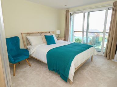A bedroom with a bed blue chair bedside table lamp and large sliding glass door at Manhattan Beach in Castletown Portland