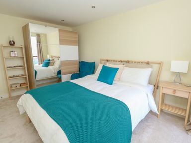 A bedroom with a bed with teal pillows and blanket a wooden wardrobe with mirrored doors a blue armchair a wooden nightstand with a lamp and a wooden shelving unit at Manhattan Beach in Castletown Portland