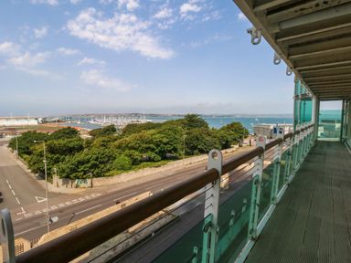A balcony with glass railing overlooking a road trees a marina with boats and a body of water at Manhattan Beach in Castletown Portland