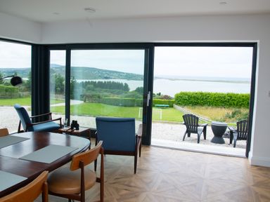 A dining area with wooden chairs and table next to a living space with blue armchairs and a floor lamp overlooking a garden and water at Ballyness Bay in Killult Upper near Gortahork County Donegal