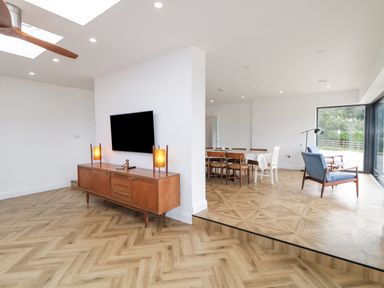 An open living and dining room with a TV mounted on the wall above a wooden cabinet a dining table with chairs and two blue armchairs near a large window at Ballyness Bay in Killult Upper near Gortahork County Donegal