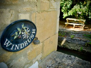 A stone wall with a decorative sign saying Windrush and a small bell next to a stone ledge and a wooden bench under trees at Windrush in Burford