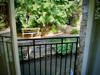 A view from a balcony overlooking a small river with stone walls and a wooden picnic table surrounded by trees at Windrush in Burford