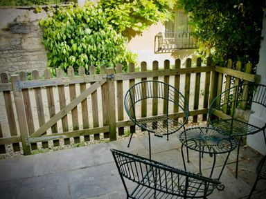 An outdoor patio with metal chairs and table behind a wooden fence with greenery in the background at Windrush in Burford