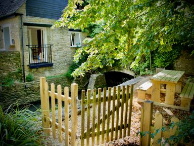 A wooden gate in front of a stone house with an outdoor picnic table and benches under a tree at Windrush in Burford