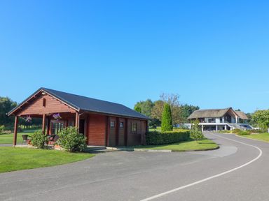 A small wooden cabin with chairs and hanging plants next to a curved driveway and a larger building with a thatched roof at An Teach Ban in Bunclody County Wexford