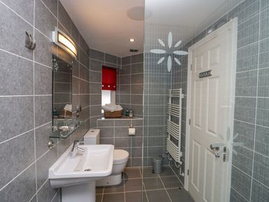 A bathroom with a sink, toilet, glass shower screen, towel radiator, and a window with a red blind at Ramblers Lodge in Bowland Bridge near Bowness-On-Windermere Lake District