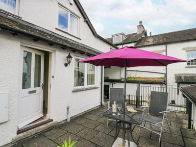 An outdoor patio area with a table, two chairs, a purple umbrella, and a white door at Ramblers Lodge Bowland Bridge near Bowness-On-Windermere Lake District