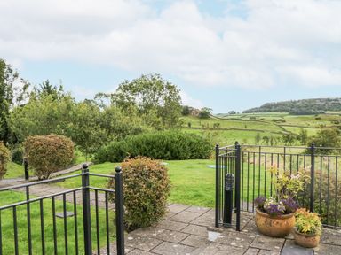 A garden with bushes, flower pots, a metal fence, and a view of green fields and hills at Ramblers Lodge in Bowland Bridge near Bowness-On-Windermere Lake District