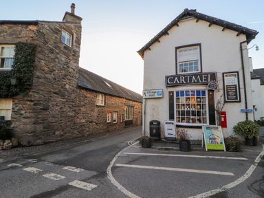 A street corner with stone buildings and a shop named Cartmel village shop with signs and planters at Ramblers Lodge Bowland Bridge near Bowness-On-Windermere Lake District