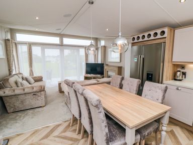 A dining area with a wooden table and six chairs next to a kitchen with a fridge and kettle and a living area with sofas and a TV at Finch 65 in Cayton Bay