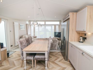 A dining table with chairs next to a kitchen area with a refrigerator and a kettle in a living space with sofas and a TV at Finch 65 in Cayton Bay