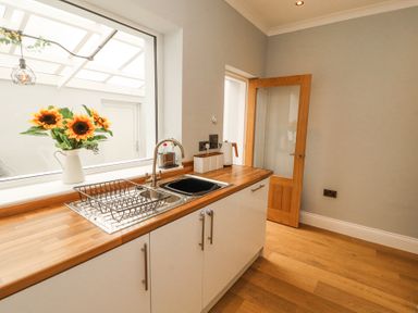 A kitchen with a sink a drying rack a vase of sunflowers on the window sill and a wooden door at Grace House in Silloth