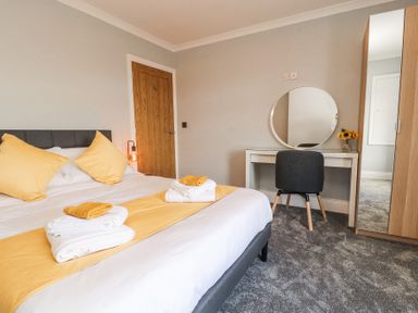 A bedroom with a bed with yellow pillows and towels a dressing table with a round mirror and a chair at Grace House in Silloth