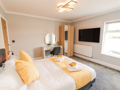 A bedroom with a double bed with yellow pillows and a runner a wardrobe a dressing table a chair and a wall mounted tv at Grace House in Silloth