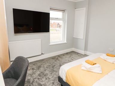 A bedroom with a bed with folded towels on top a grey chair a wall-mounted TV a radiator and a window at Grace House in Silloth
