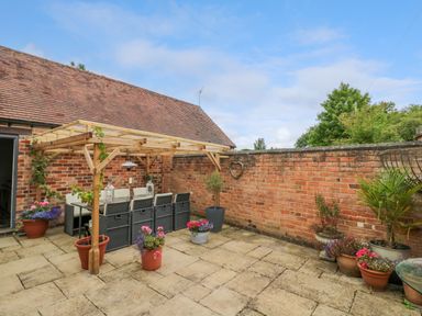 An outdoor patio area with a wooden pergola covering a dining table and chairs surrounded by potted plants and a brick wall at The Bit on the Side in Ettington