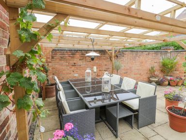 An outdoor patio dining area with a glass table and cushioned chairs under a wooden pergola surrounded by potted plants and flowers at The Bit on the Side in Ettington