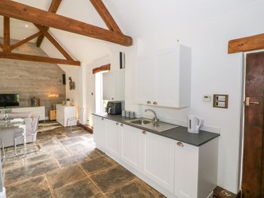 A kitchen with white cabinets a sink and kettle next to a dining area with a glass table and chairs and wooden beams on the ceiling at The Bit on the Side in Ettington