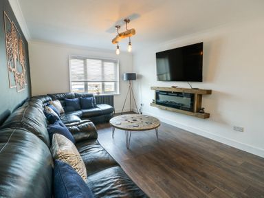 A living room with a black leather corner sofa a round wooden coffee table a wall-mounted TV and an electric fireplace at Ty Copr in Amlwch