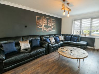 A living room with a black leather sectional sofa with pillows and a wooden round coffee table at Ty Copr in Amlwch