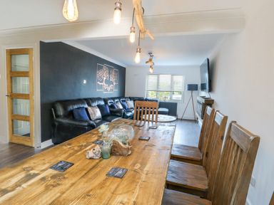 A living room with wooden dining table and chairs a black leather sofa and wall-mounted tv at Ty Copr in Amlwch