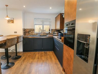 A kitchen with a wooden floor, black lower cabinets, wooden upper cabinets, a built-in oven, refrigerator, sink under a window, and a breakfast bar with stools at Ty Copr in Amlwch