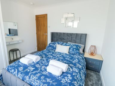 A bedroom with a bed with blue floral bedding and rolled white towels on it a wooden door a dark bedside table with a lamp and a small vanity with a stool at Ty Copr in Amlwch