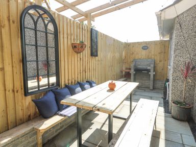 An outdoor patio area with wooden benches and table along a wooden fence with cushions and a wall mirror and a barbecue grill and potted plants at Ty Copr in Amlwch