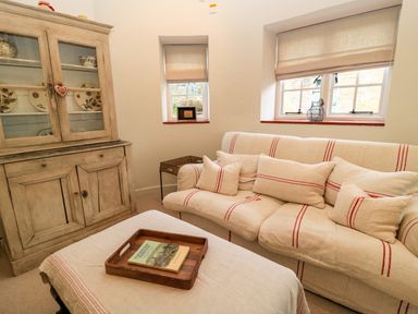 A sitting room with sofa, cushions, wooden cabinet, and ottoman with tray and book at The Elm Coach House in Chipping Norton