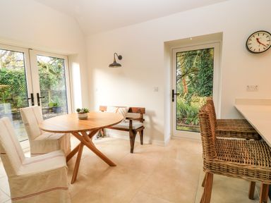 A dining area with a wooden round table two fabric chairs a wooden bench and wicker stools at The Elm Coach House in Chipping Norton
