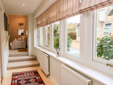 A hallway with stairs leading to a sitting area and large windows with striped blinds at The Elm Coach House in Chipping Norton